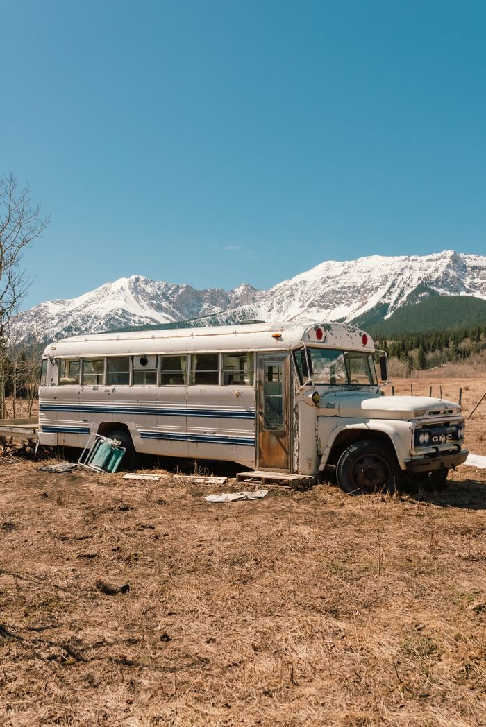 An old bus sits abandoned in a desert field with snow-capped mountains in the background.