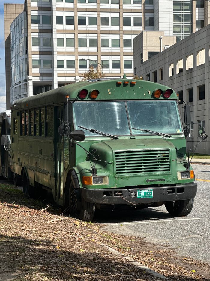 Old green bus parked on a city street with buildings in the background.